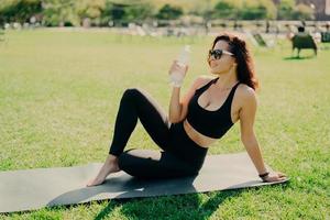 photo extérieure d'une femme mince active boit de l'eau de la bouteille a soif après un entraînement de fitness concentré sur la distance porte un haut court et des leggings pratique le yoga à l'extérieur. notion de sport