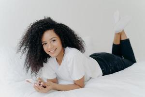 photo intérieure d'une femme joyeuse du millénaire avec une coiffure afro, allongée sur le ventre dans un lit confortable, discutant en ligne, vérifiant la boîte e-mail, portant un t-shirt blanc, des chaussettes et un pantalon noir, a un après-midi paresseux