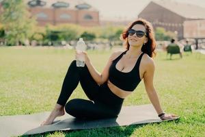 une femme active positive a une forme de corps parfaite grâce à des poses d'entraînement physique régulières sur karemat avec une bouteille d'eau se repose après l'entraînement passe du temps libre à l'extérieur. pratiquer le yoga le matin photo