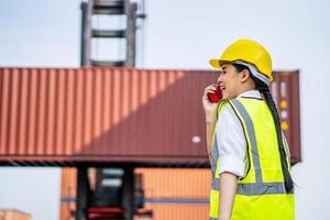 jeune femme ingénieur confiant souriant et utilisant la communication radio et portant un casque de sécurité jaune et vérifier le contrôle de la boîte de conteneurs de chargement du navire de fret pour l'importation et l'exportation photo