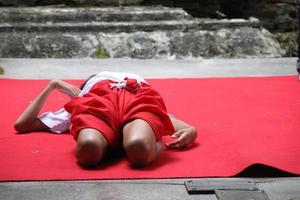 un enfant pratique son corps sur un tapis rouge au sol en ciment pour apprendre khon, bangkok. khon est un genre dramatique de danse en thaïlande. photo