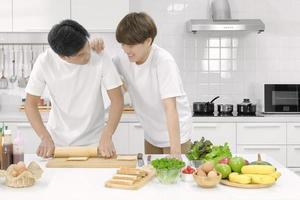 jeune couple lgbt masculin asiatique heureux de passer du temps ensemble pendant la cuisson de la salade de pain dans une cuisine blanche avec un visage souriant. mise au point sélective. photo