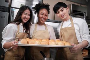 portrait de trois partenaires de démarrage de pâtisserie regardant la caméra avec un sourire joyeux et fier avec un plateau de pain dans la cuisine. un ami et fondateur d'aliments de boulangerie et de petite entreprise de boulangerie fraîche quotidienne. photo
