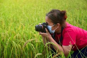 un senior asiatique porte un jean, un t-shirt rouge, un masque et tient un appareil photo numérique pour filmer joyeusement les épis de riz dans la rizière verte. mise au point douce et sélective, vie heureuse après le concept de retraite anticipée.