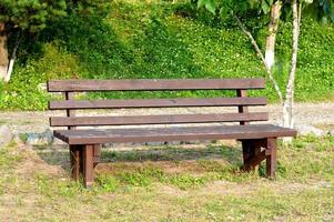 banc en bois marron dans le parc. journée d'été ensoleillée. herbe verte et arbres. espace de repos et de détente. banc vide pour s'asseoir. matériau extérieur en bois. photo