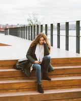 belle femme aux cheveux longs est assise dans les escaliers avec un téléphone à la main, dos à la caméra. automne ou hiver, fille en veste à l'extérieur en attente d'appel ou de sms, textos photo