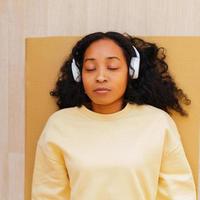femme afro-américaine dans les écouteurs se reposant sur un tapis avec les yeux fermés. se détendre après l'exercice