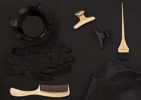 accessoires pour la coloration des cheveux sur fond sombre. un peigne, une brosse, un bol, des pinces et une cape sont disposés sur la table. articles de coiffeurs en noir et or, mise à plat inhabituelle photo