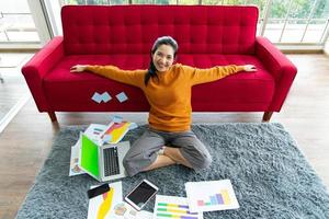 travail à domicile. jeune belle femme asiatique utilisant un ordinateur portable avec un gadget assis sur un tapis gris à la maison. photo