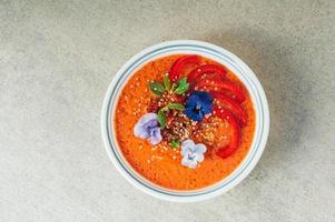savoureuse soupe de tomates dans un bol blanc, décorée de petites fleurs, de poivron rouge, de graines de chanvre et de menthe. repas délicieux. concept de plat végétarien. l'heure du dîner photo
