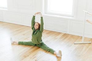 une femme aux cheveux rouges et bouclés, forte et flexible, se fend la jambe latérale, lève les mains, veut avoir un corps parfait, porte des vêtements de sport, s'entraîne dans un studio de ballet. danseur talentueux a répétition intérieure photo