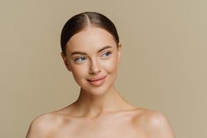 portrait d'une belle jeune femme à la peau propre porte un maquillage minimal avec une expression rêveuse pose nue à l'intérieur isolée sur fond marron. concept de beauté et de cosmétologie féminine. photo