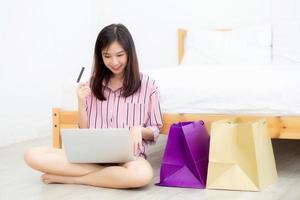 belle femme asiatique shopping en ligne avec ordinateur portable assis sur le sol dans la chambre, fille tenant l'achat par carte de crédit et sacs à provisions, concept de style de vie. photo