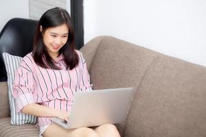 belle de portrait jeune femme asiatique travaillant sur un ordinateur portable en ligne avec sourire et heureux assis sur un canapé dans le salon, fille utilisant un ordinateur portable avec connexion à internet, concept d'entreprise et de réussite. photo