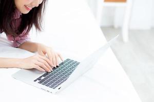 belle jeune femme asiatique allongée sur le lit à l'aide d'un ordinateur portable dans la chambre pour les loisirs et la détente, indépendante avec un cahier de travail fille, concept de communication. photo