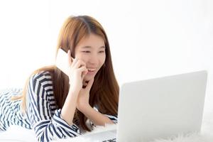 belle jeune femme asiatique allongée sur le lit à l'aide d'un ordinateur portable et parlant d'un téléphone intelligent mobile dans la chambre pour les loisirs et la détente, indépendante avec un cahier de travail pour fille, concept de communication. photo