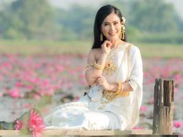 une élégante femme thaïlandaise portant des vêtements traditionnels thaïlandais ornés d'ornements d'or, assise sur un pont en bois dans un champ de lotus photo