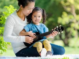 une fille asiatique est assise sur le tapis avec sa mère et sa mère lui apprend à jouer du ukulélé photo