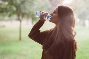 jeune belle fille buvant de l'eau d'une bouteille en plastique dans la rue dans le parc en automne ou en hiver. une femme avec de beaux longs cheveux noirs et épais étanche sa soif d'eau lors d'une promenade photo