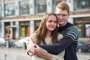 portrait d'un couple heureux embrassant au centre-ville, homme aux cheveux roux avec des lunettes regardant droit, femme aux cheveux longs regardant loin photo