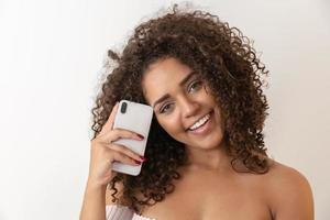 prise de vue en studio d'une jolie fille afro-américaine heureuse et divertie avec une coiffure afro tenant un smartphone à l'aide d'un appareil pour s'amuser photo