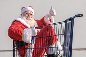 chariot de supermarché avec le père noël à l'intérieur. Pouce en l'air. shopping pour le concept de noël. photo