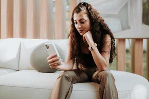 heureuse dame latine se détendre seule à la maison s'asseoir sur la pose de la chambre partager de bonnes nouvelles sur les réseaux sociaux via un téléphone portable. photo