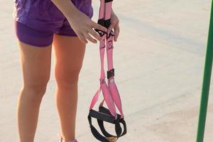 belle femme en forme de sportwear rose et violet s'entraînant sur une salle de sport en plein air le matin, exercices avec des sangles de suspension dans le parc photo