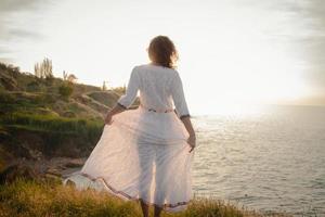 jeune femme marchant sur la plage du matin dans une belle robe blanche. femme en forme ayant du bon temps pendant le lever du soleil. photo