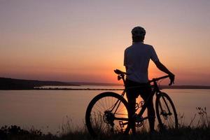 silhouette d'un cycliste regardant le coucher du soleil dans le lac, cycliste masculin en casque pendant le coucher du soleil photo