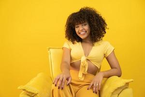 heureuse femme africaine américaine qui rit avec ses cheveux bouclés sur fond jaune. femme bouclée riante en pull touchant ses cheveux et regardant la caméra. photo