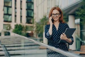 femme rousse en vêtements à la mode a une conversation téléphonique marche dans la rue près d'un bâtiment moderne photo