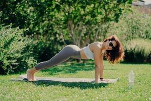 concept de mode de vie sain. photo horizontale d'une femme de fitness motivée en vêtements de sport pose en pose de planche sur des exercices de tapis de fitness à l'extérieur bénéficie d'un entraînement régulier porte des lunettes de soleil à l'heure d'été