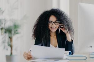 une chef de la direction expérimentée a une conversation téléphonique, résout un problème, se concentre sur des documents, porte des lunettes et des vêtements de cérémonie, pose sur le lieu de travail, a une expression joyeuse. directeur administratif photo