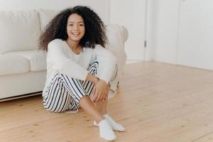 photo intérieure d'une femme afro-américaine heureuse et reposante assise sur le sol dans le salon, un canapé blanc en arrière-plan, porte des vêtements et des chaussettes à la mode, exprime des émotions positives, se repose seule à la maison