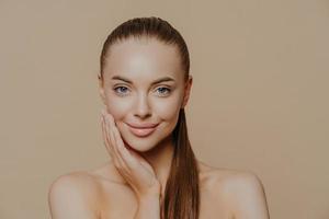 photo d'une belle jeune femme a une peau lisse et saine après des procédures de beauté et reçoit un massage facial, se tient les épaules nues, a de longs cheveux raides et foncés, isolée sur un mur beige se soucie de son corps