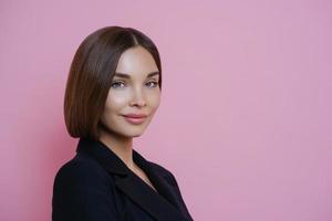 photo de profil d'une femme aux cheveux noirs vêtue d'un costume noir, a un maquillage naturel, a l'air sûre d'elle à l'intérieur, pose sur fond rose, espace vide pour votre promotion. concept de femmes et d'élégance