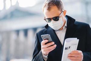 photo en extérieur d'un directeur d'homme sérieux concentré sur l'affichage cellulaire, lit les nouvelles en ligne, tient un journal, porte un masque médical pour se protéger contre le coronavirus, l'infection par le virus de la grippe, pose dans la rue