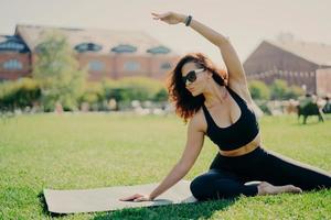 image d'une femme en forme motivée lève le bras et fait des exercices d'étirement à l'extérieur pose sur un tapis de fitness porte des lunettes de soleil et des vêtements de sport s'entraîne activement. entraîneur de fitness fait du sport en plein air photo