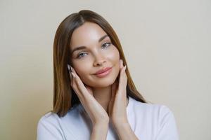 beauté naturelle et concept de femmes. gros plan d'une jolie jeune femme charmante touche doucement les joues, jouit d'une peau saine, habillée avec désinvolture, a de longs cheveux bien soignés, porte du maquillage, pose à l'intérieur photo