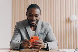 jeune entrepreneur africain souriant assis au bureau et utilisant un téléphone portable sur le lieu de travail photo