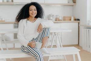 une femme afro-américaine heureuse tient une tasse de boisson chaude, se penche au genou, porte un pull blanc élégant et un pantalon rayé, sourit agréablement, passe du temps libre à la maison, s'assied sur un banc dans la cuisine photo