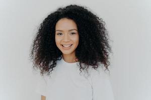 concept de bonheur, de bien-être et d'émotions positives. photo de tête d'une femme à l'air agréable avec des cheveux croustillants, des sourires et des rires, a une peau saine, porte un t-shirt décontracté, des modèles contre le mur blanc du studio.