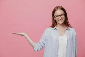 photo horizontale d'une jeune femme séduisante détient un espace libre, vêtue de vêtements formels, sourit doucement, pose sur fond rose, annonce un nouvel article dans la boutique, a une expression faciale heureuse. publicité