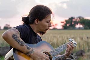 jeune homme tatoué jouer à la guitare acoustique dans les champs d'été pendant le beau coucher de soleil photo