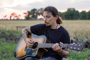 jeune homme tatoué jouer à la guitare acoustique dans les champs d'été pendant le beau coucher de soleil photo