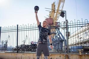 jeune athlète masculin barbu s'entraînant dans une zone industrielle par temps ensoleillé, exercices de kettlebells à l'extérieur, arrière-plan urbain photo