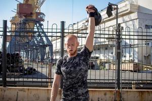 jeune athlète masculin barbu s'entraînant dans une zone industrielle par temps ensoleillé, exercices de kettlebells à l'extérieur, arrière-plan urbain photo