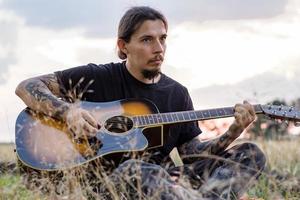 jeune homme tatoué jouer à la guitare acoustique dans les champs d'été pendant le beau coucher de soleil photo