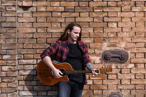 jeune homme aux cheveux longs et tatouage jouer à la guitare acoustique à l'extérieur dans la rue photo